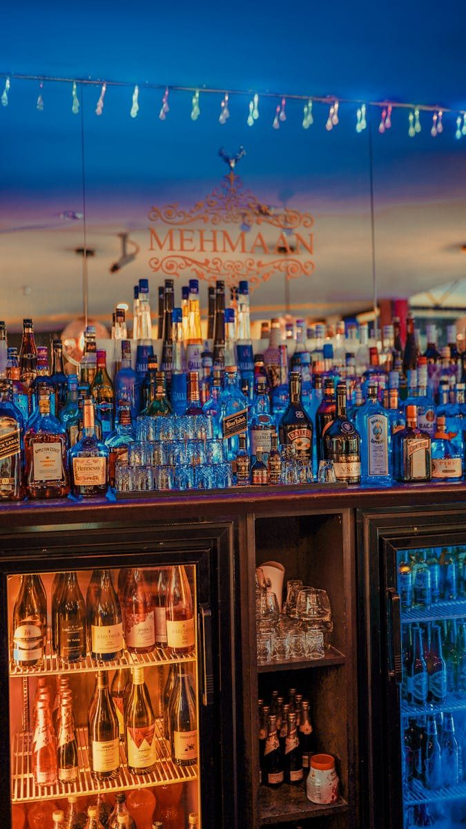 Colorful bar setup with a variety of bottles, a mirror backdrop, and a refrigerator.